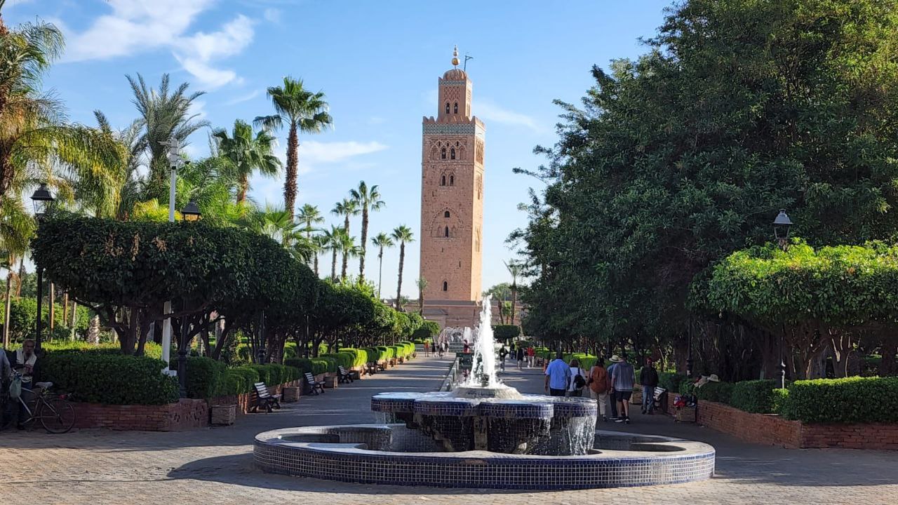 Koutoubia Mosque Minaret from Lalla Hasna Park, Marrakech - Private Tours of Morocco - Private Tours of Morocco A scenic view of the tall sandstone Koutoubia Mosque minaret towering over Lalla Hasna Park in Marrakech, featuring a tiled water fountain in the foreground and a tree-lined walkway with palm trees under a bright blue sky.