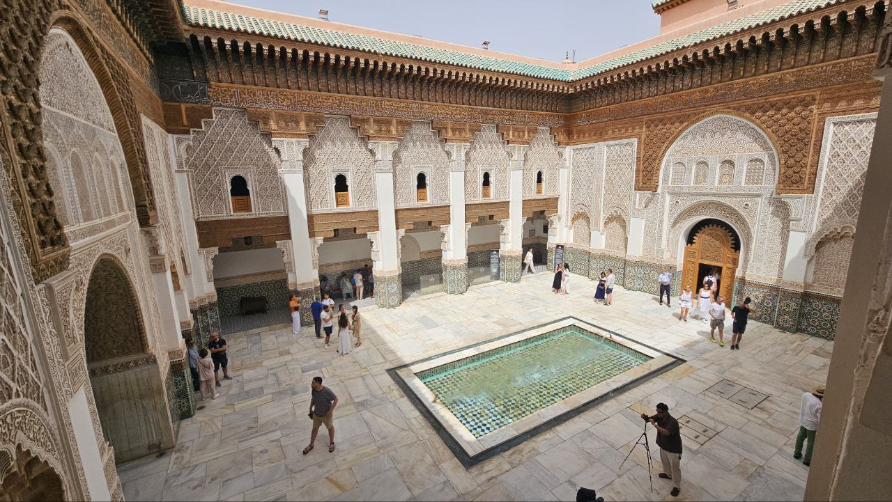 High-angle view of the central marble courtyard at Ben Youssef Madrasa in Marrakech, showing a rectangular reflection pool surrounded by tourists and towering walls adorned with detailed Islamic geometric carvings, stucco work, and dark cedar wood trim.